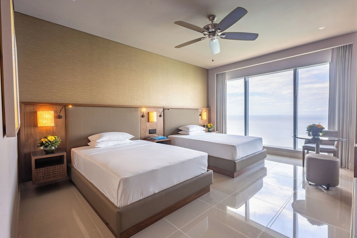 A hotel room with two beds, a ceiling fan, a large window with a view, and a small table with a chair.