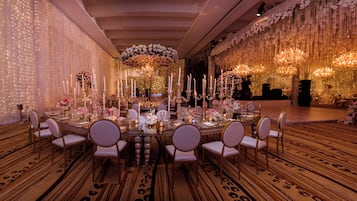 Uma sala de banquetes com uma longa mesa posta para um evento formal, rodeada de cadeiras.