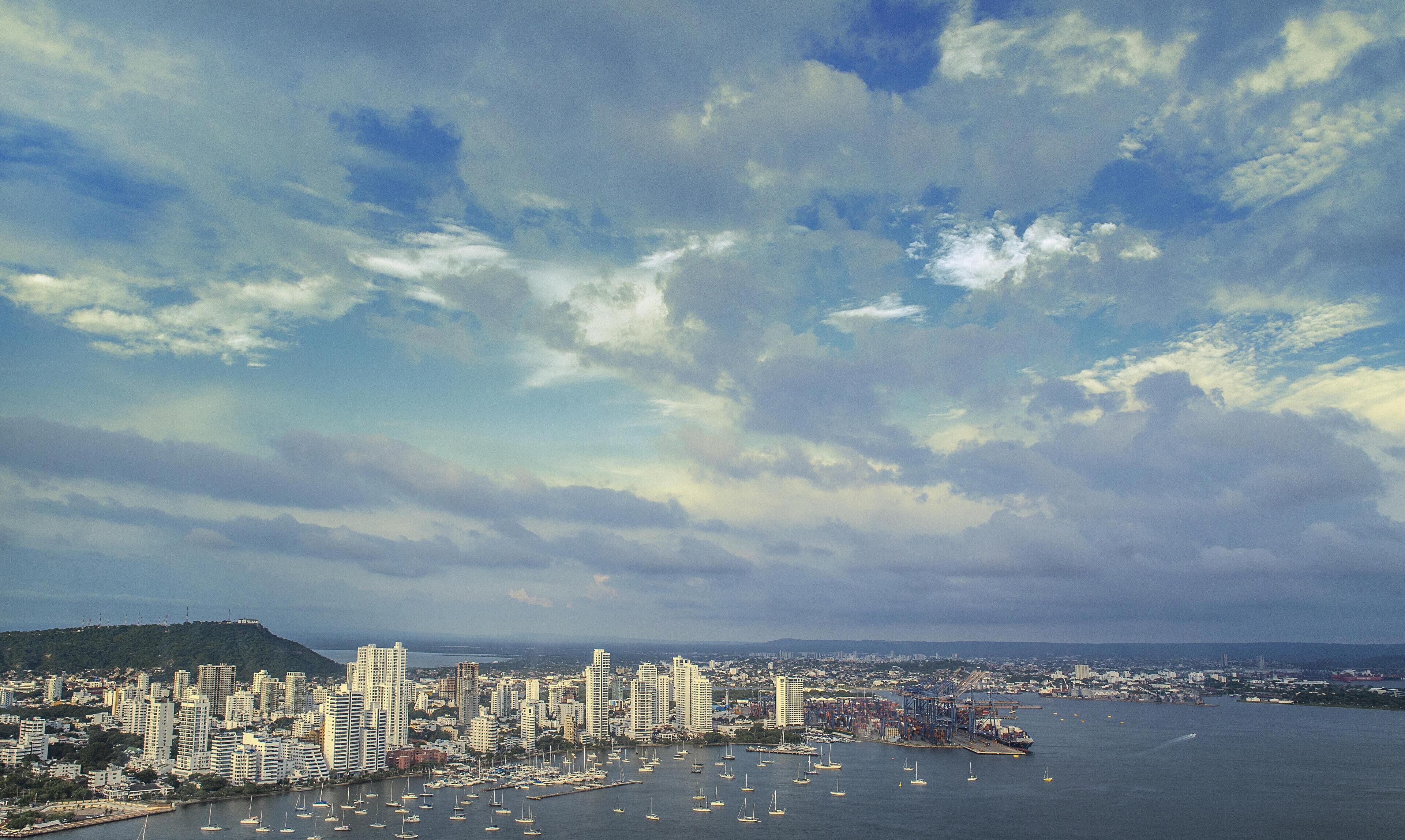 A coastal cityscape with high-rise buildings, a marina with numerous boats, and a large body of water.