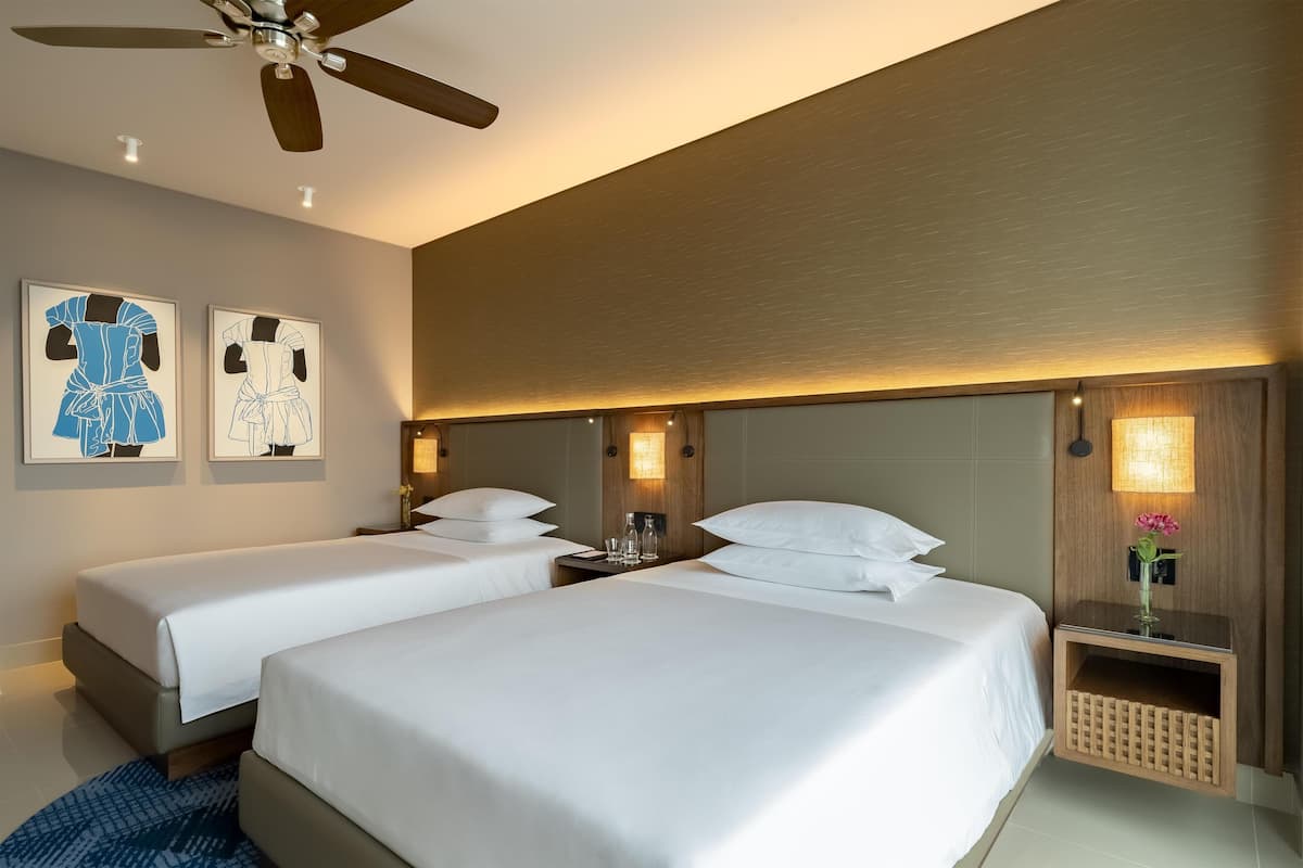 A hotel room with two single beds, a ceiling fan, wall-mounted lights, and artwork on the wall.