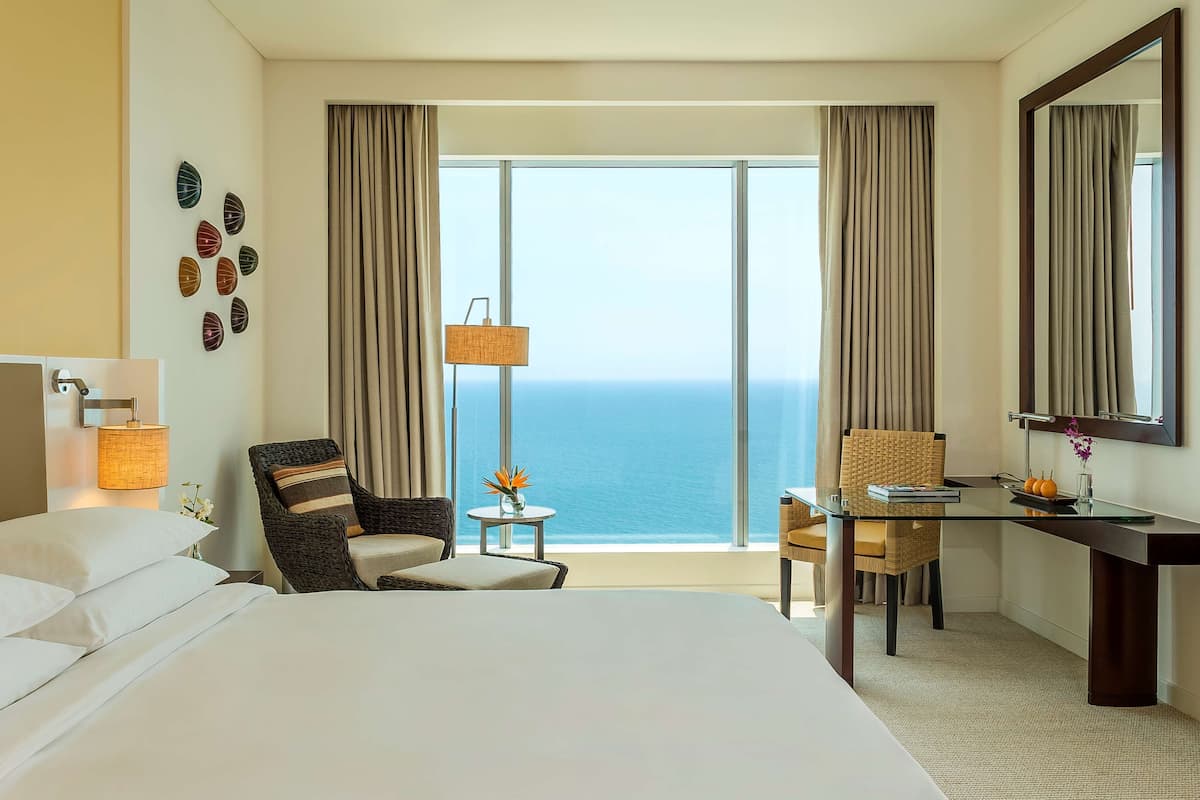 A hotel room with a bed, two chairs, a small table, a lamp, and a large window with a view of the ocean.