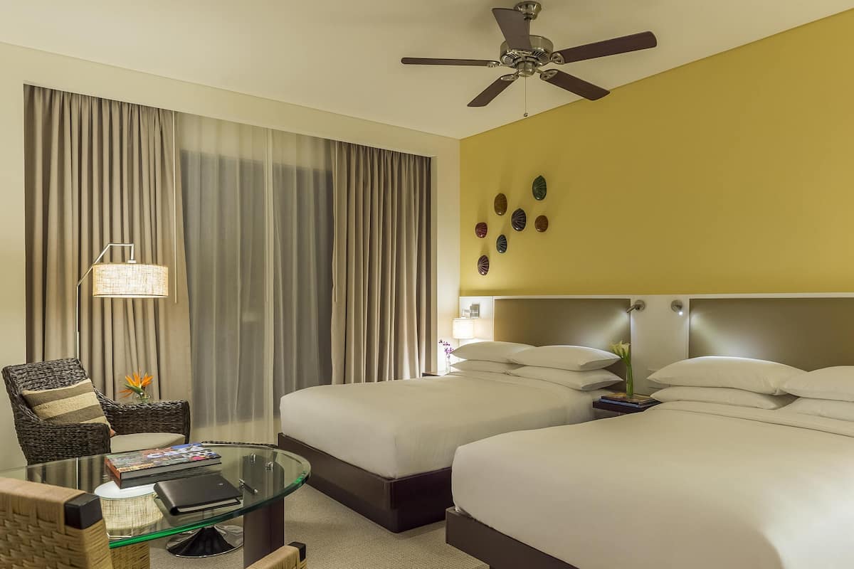 A hotel room with two beds, a glass coffee table, a wicker chair, a ceiling fan, and a decorative wall piece.