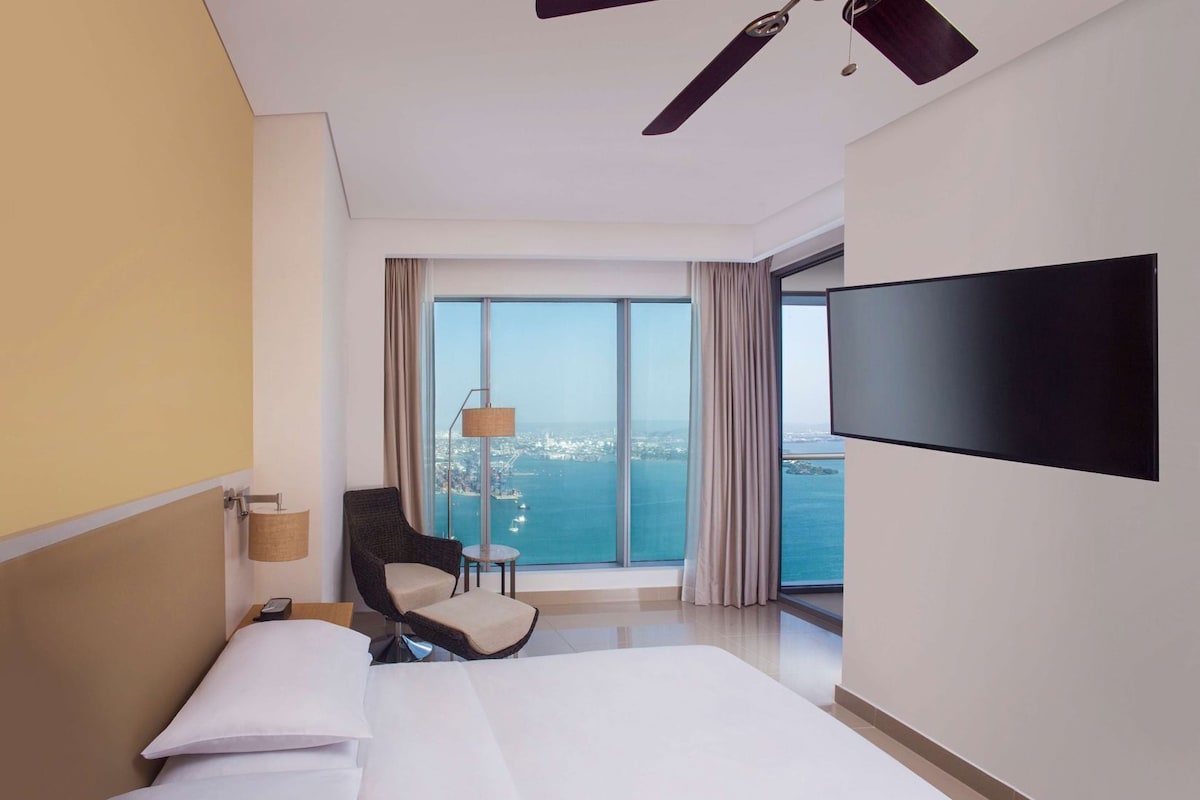 A hotel room with a bed, a chair, a TV, a lamp, and a view of the sea.