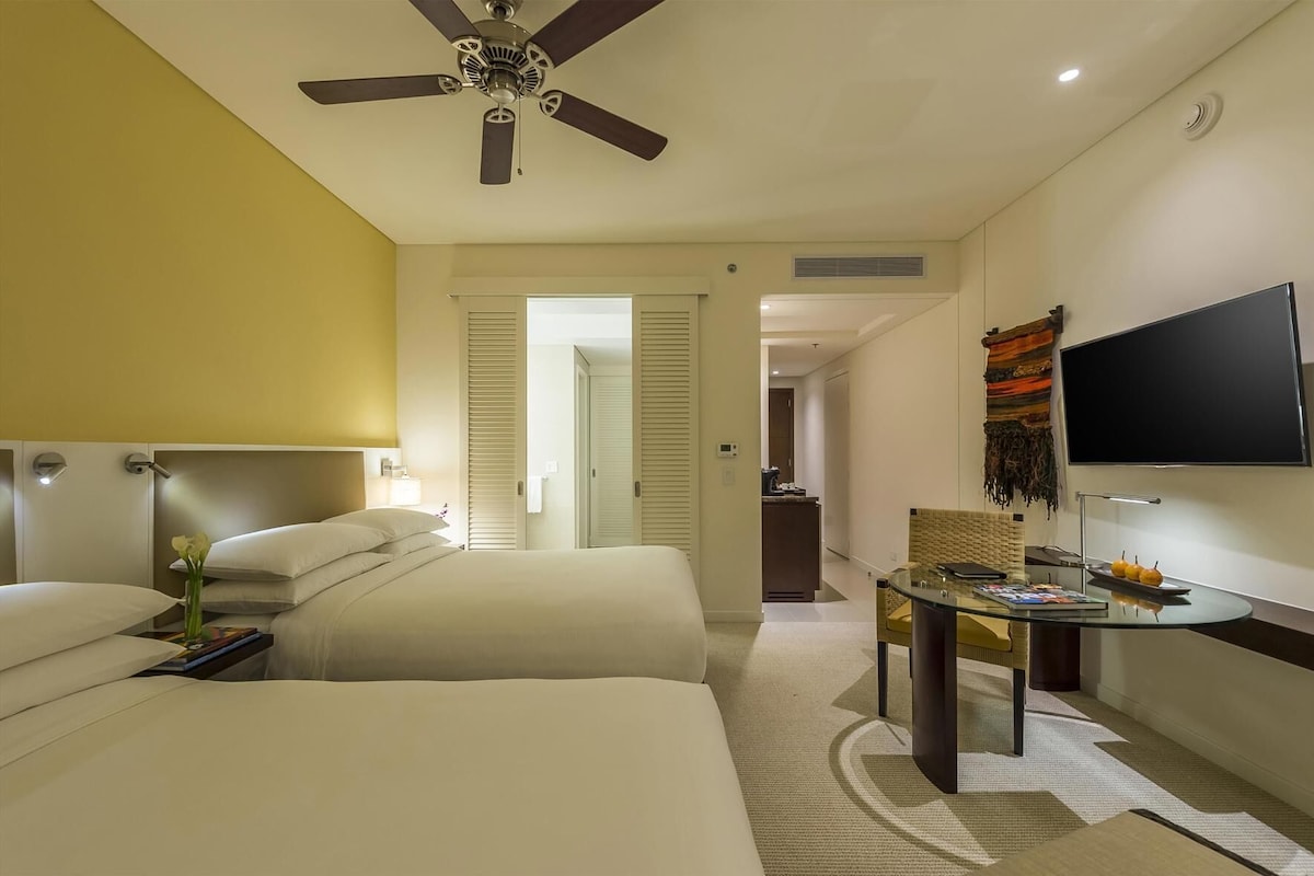 A hotel room with two beds, a ceiling fan, a TV, a small table with a magazine, and a bathroom visible through an open door.
