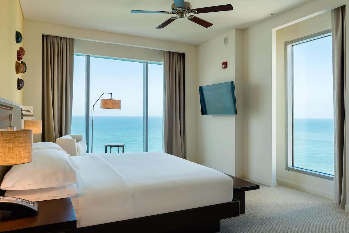 A hotel room with a large bed, a ceiling fan, a TV, and a view of the ocean.