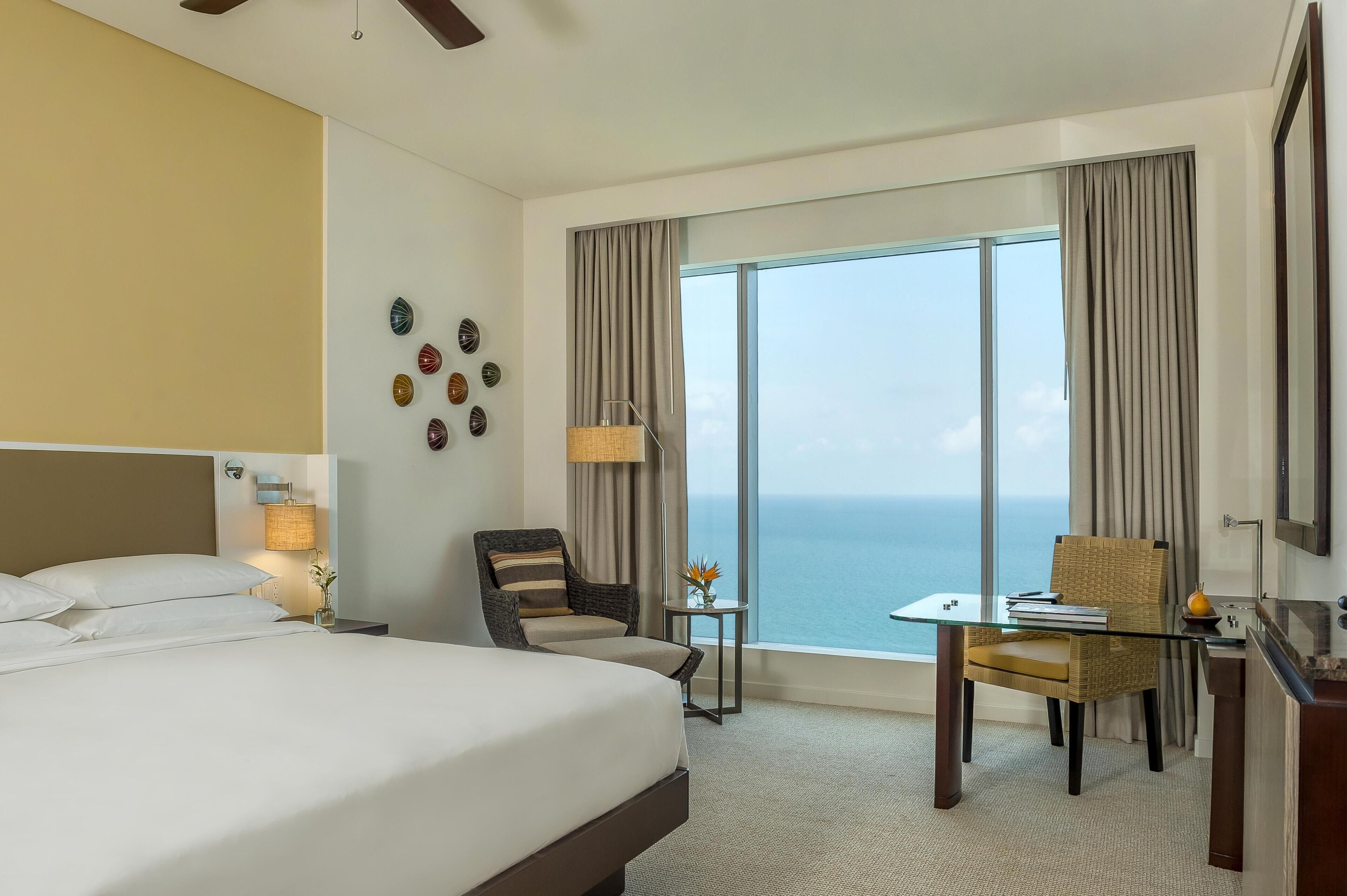 A hotel room with a large bed, a desk with a chair, a small table with a lamp, and a view of the ocean.