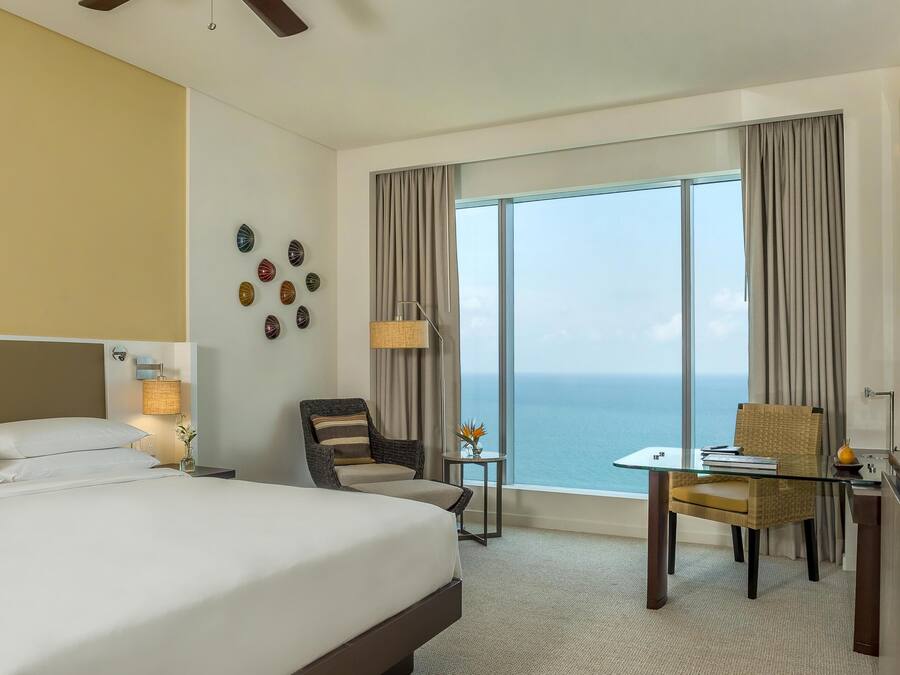 A hotel room with a large bed, a desk with a chair, a small table with a lamp, and a view of the ocean.