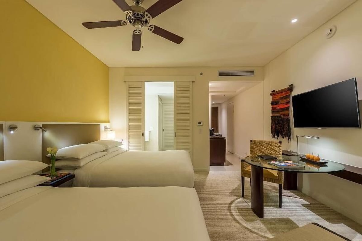 A hotel room with two beds, a ceiling fan, a TV, a desk, and a dining area.