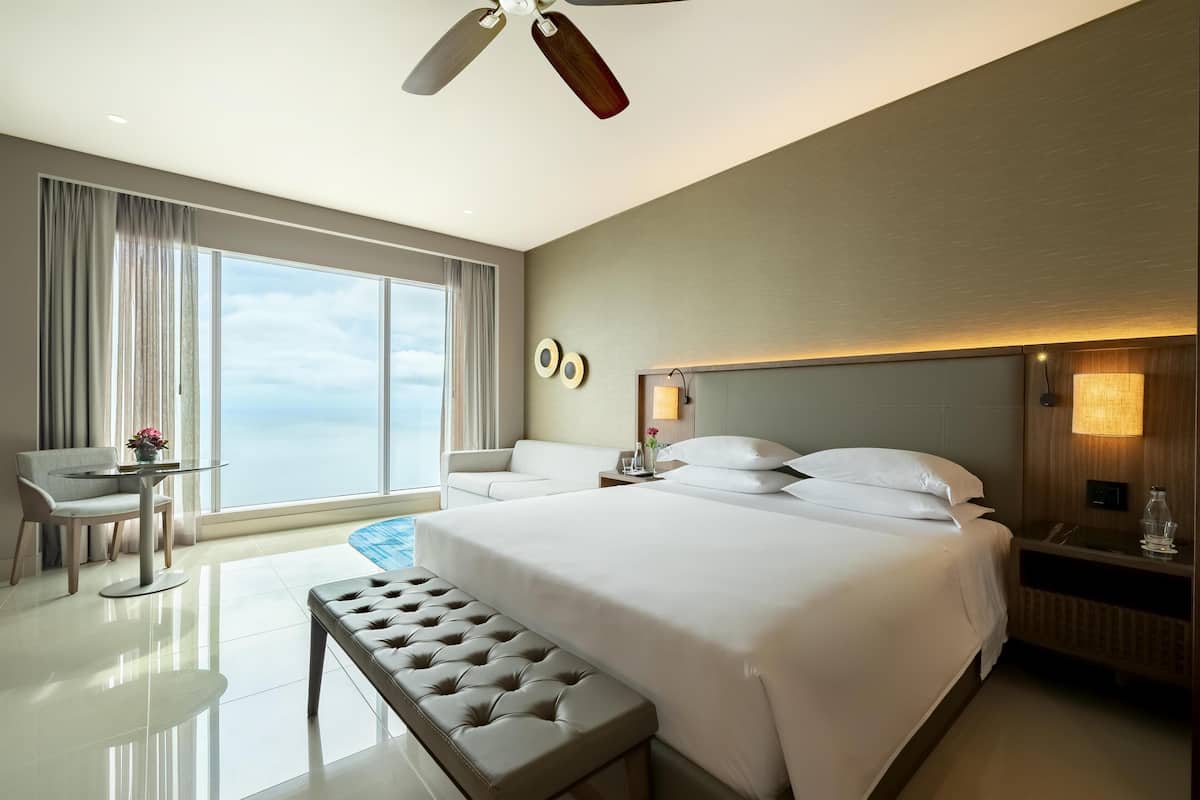 A modern hotel room with a large bed, a seating area with a table, and a view of the sky.