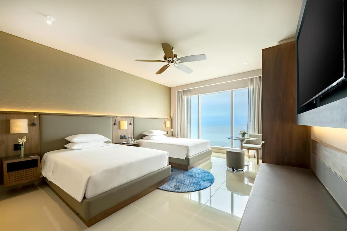 A hotel room with two beds, a ceiling fan, a TV, a desk, and a view of the ocean.