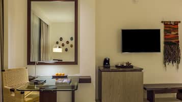 A hotel room with a glass table, wicker chair, a wooden cabinet, a flat-screen TV, and a decorative wall hanging.