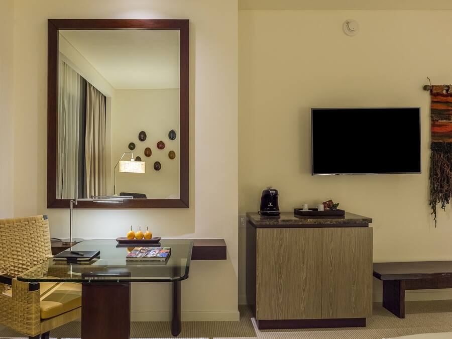 A hotel room with a glass table, wicker chair, a wooden cabinet, a flat-screen TV, and a decorative wall hanging.