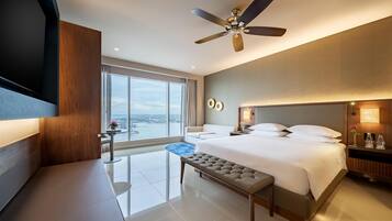 A modern hotel room with a large bed, a ceiling fan, a bench, a bedside table, and a view of the city.