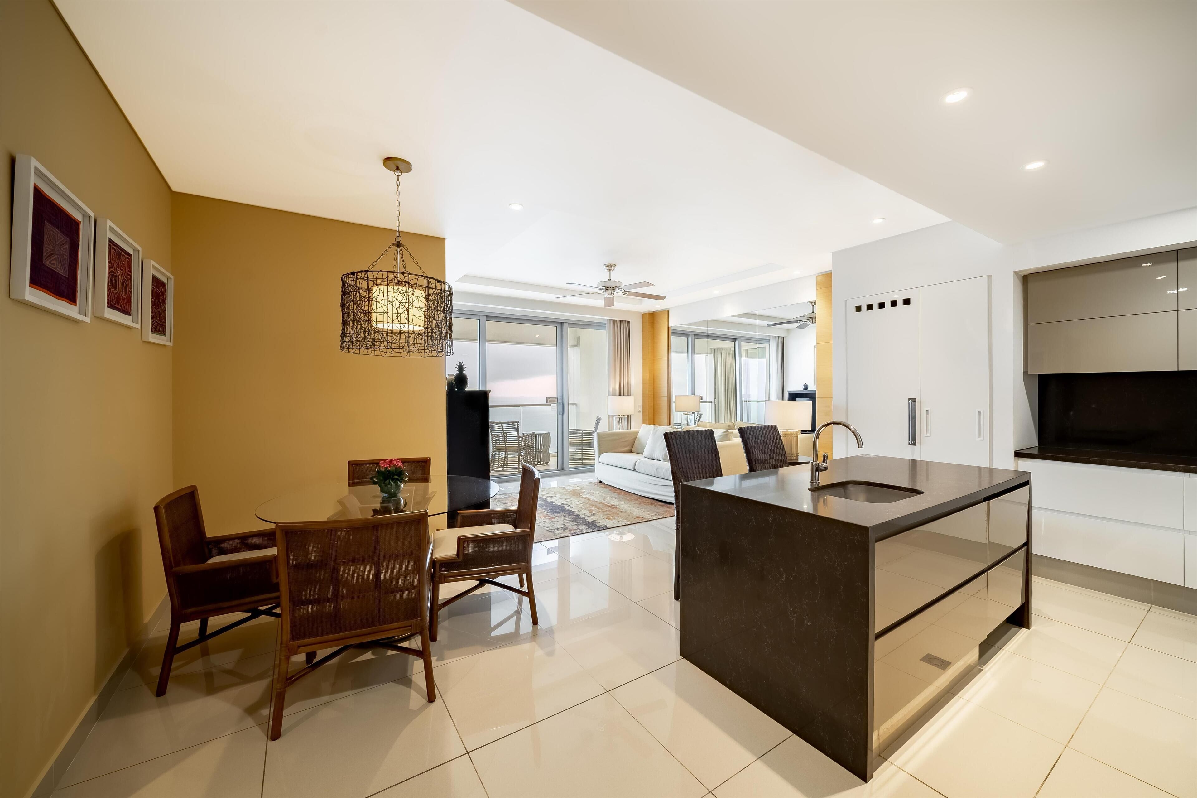 Una cocina moderna con una isla central, una zona de comedor con sillas de madera y una sala de estar con sofá y televisor.