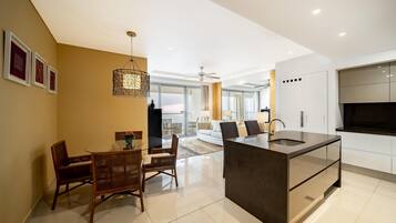 Una cocina moderna con una isla central, una zona de comedor con sillas de madera y una sala de estar con sofá y televisor.