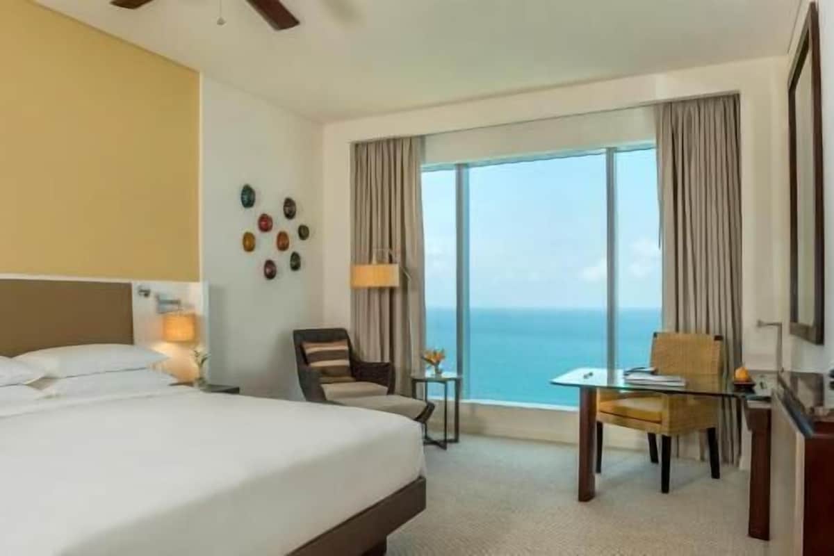 A hotel room with a bed, a desk, a chair, a view of the ocean, and a wall with decorative items.
