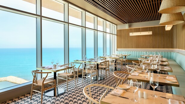 A restaurant with large windows offering a view of the sea, wooden ceiling, and modern furnishings.