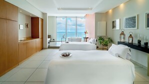 A hotel room with two beds, a view of the sea, a wooden cabinet, a small table, and a plant.