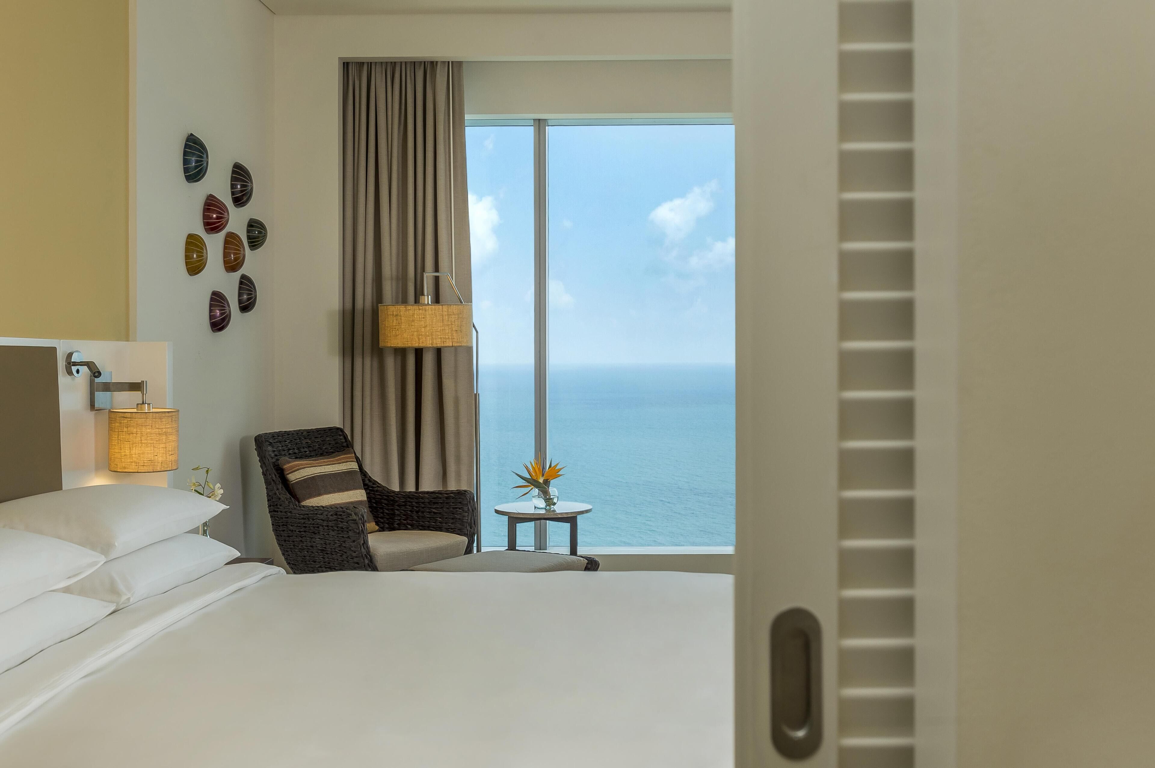 Habitación de hotel con cama, una silla, una mesita con un jarrón, una lámpara y vista al mar.