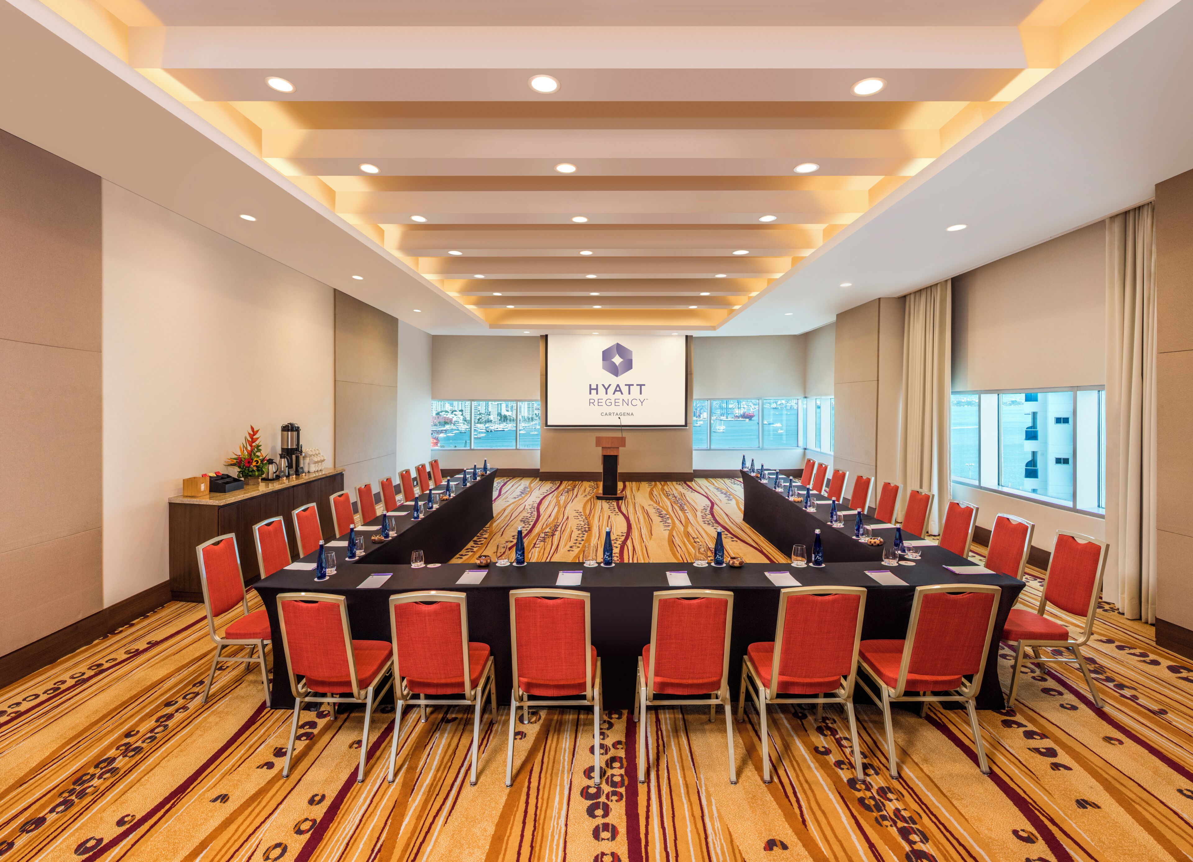 A conference room with a long table, red chairs, a podium, and a screen displaying the Hyatt Regency logo.