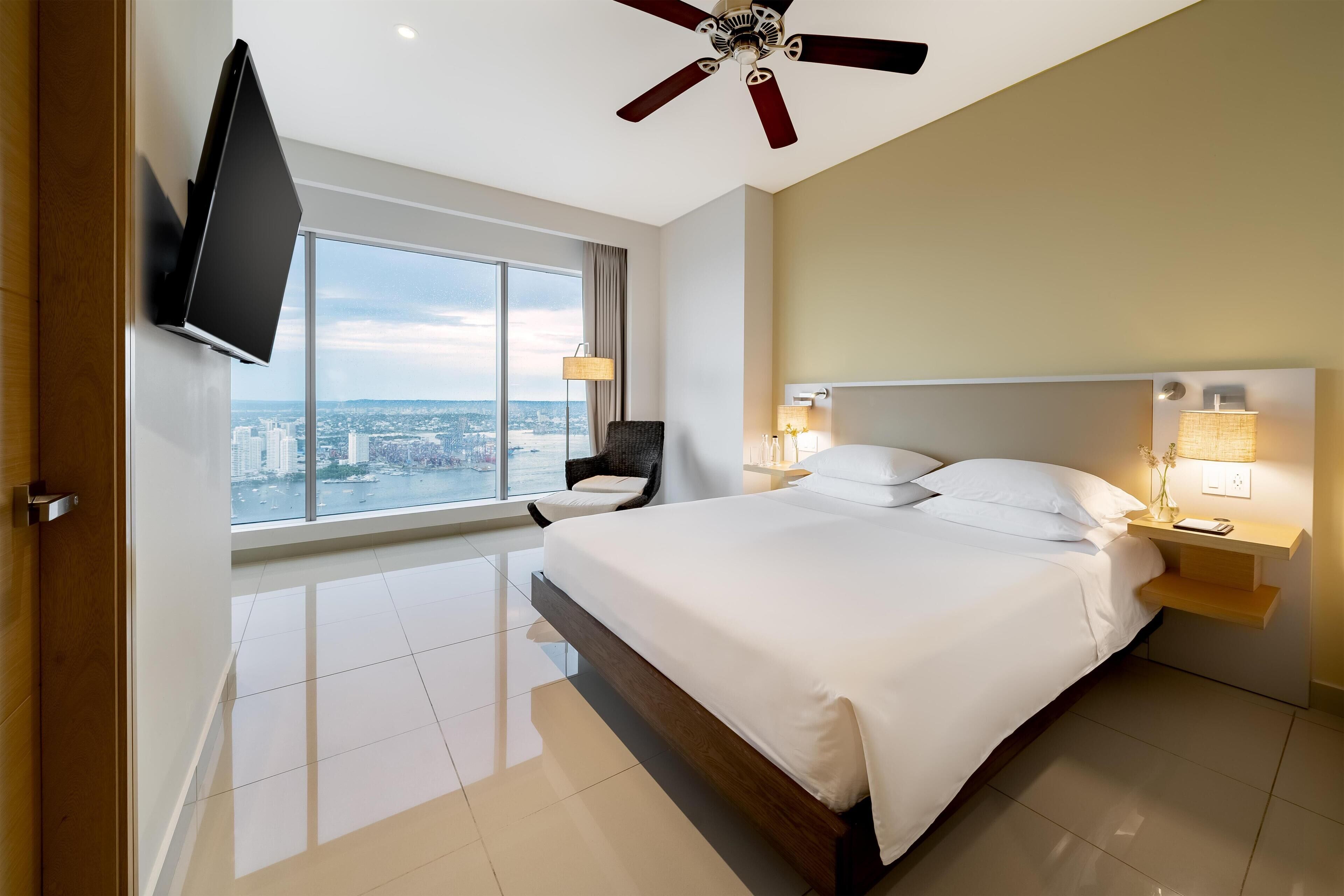 Un dormitorio de hotel moderno con una cama grande, un televisor de pantalla plana en la pared, un ventilador de techo y vistas a la ciudad desde la ventana.