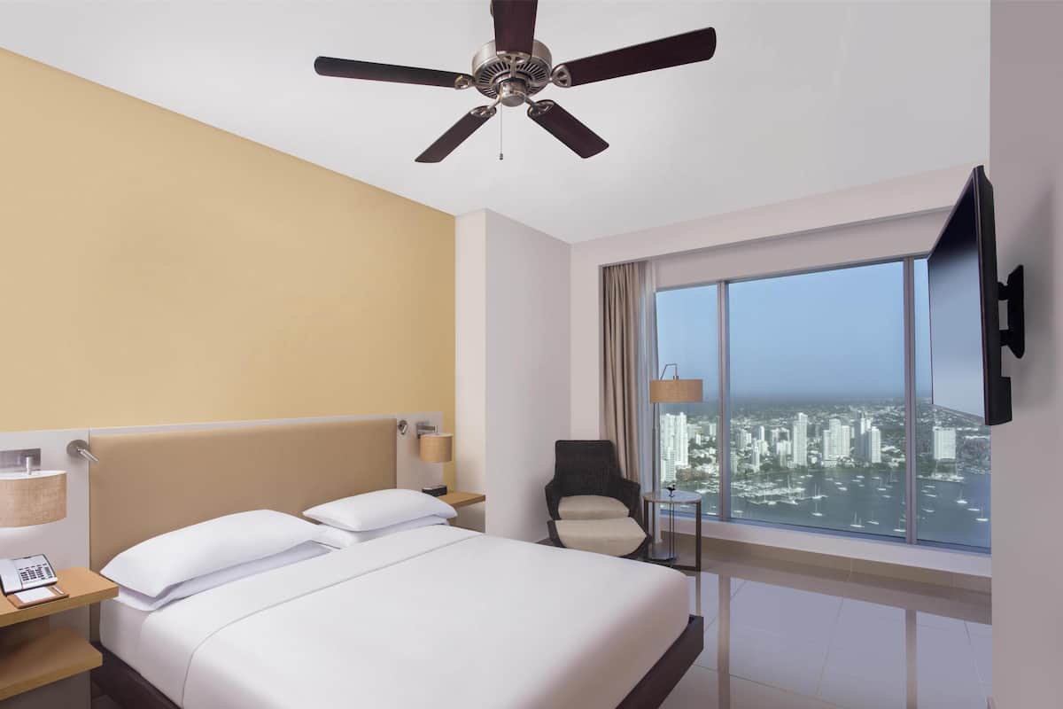 A hotel room with a large bed, a ceiling fan, a desk with a phone, a chair, a lamp, and a TV mounted on the wall.