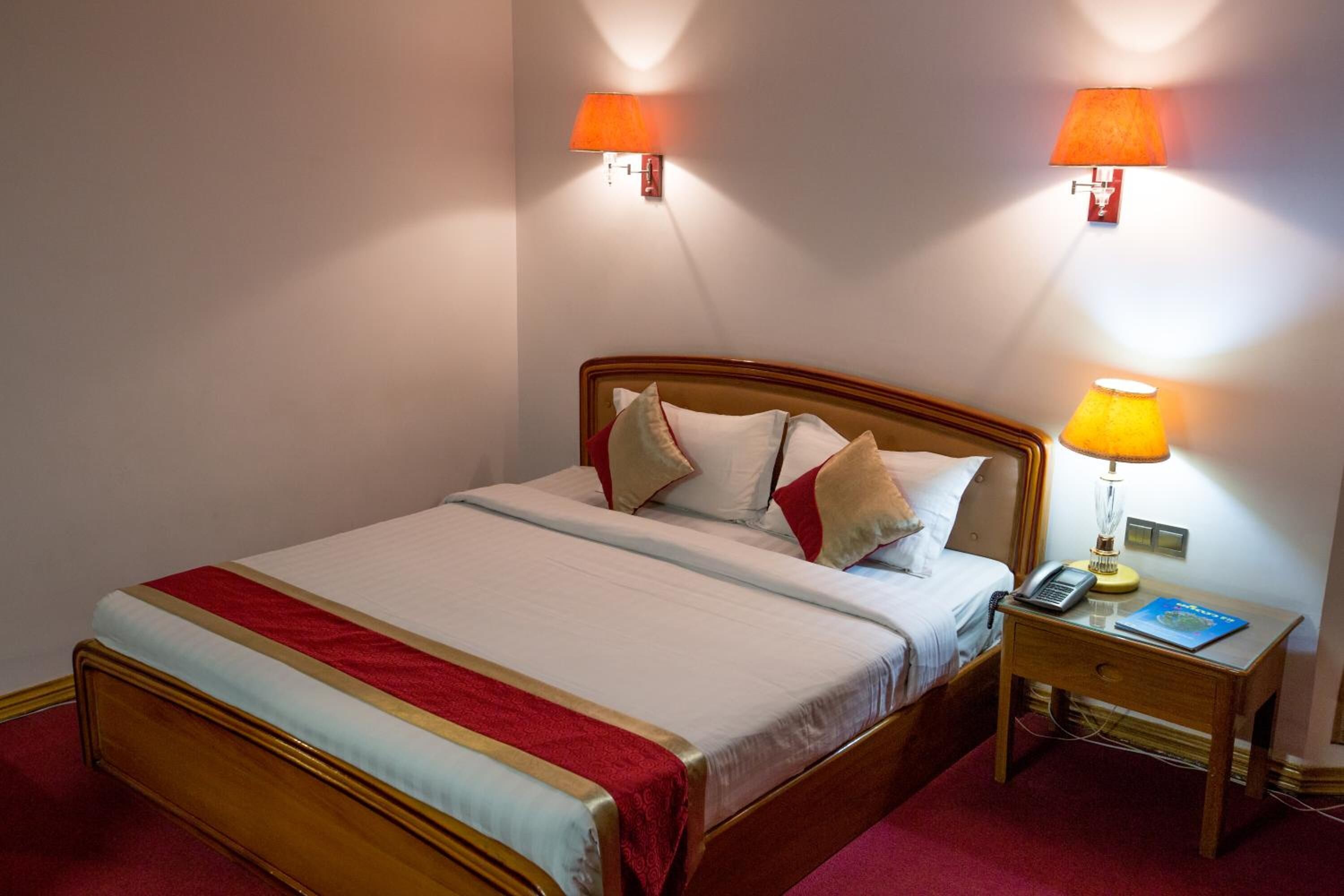 A hotel room with a wooden bed, two bedside lamps, a nightstand with a telephone and a book, and a red carpet.