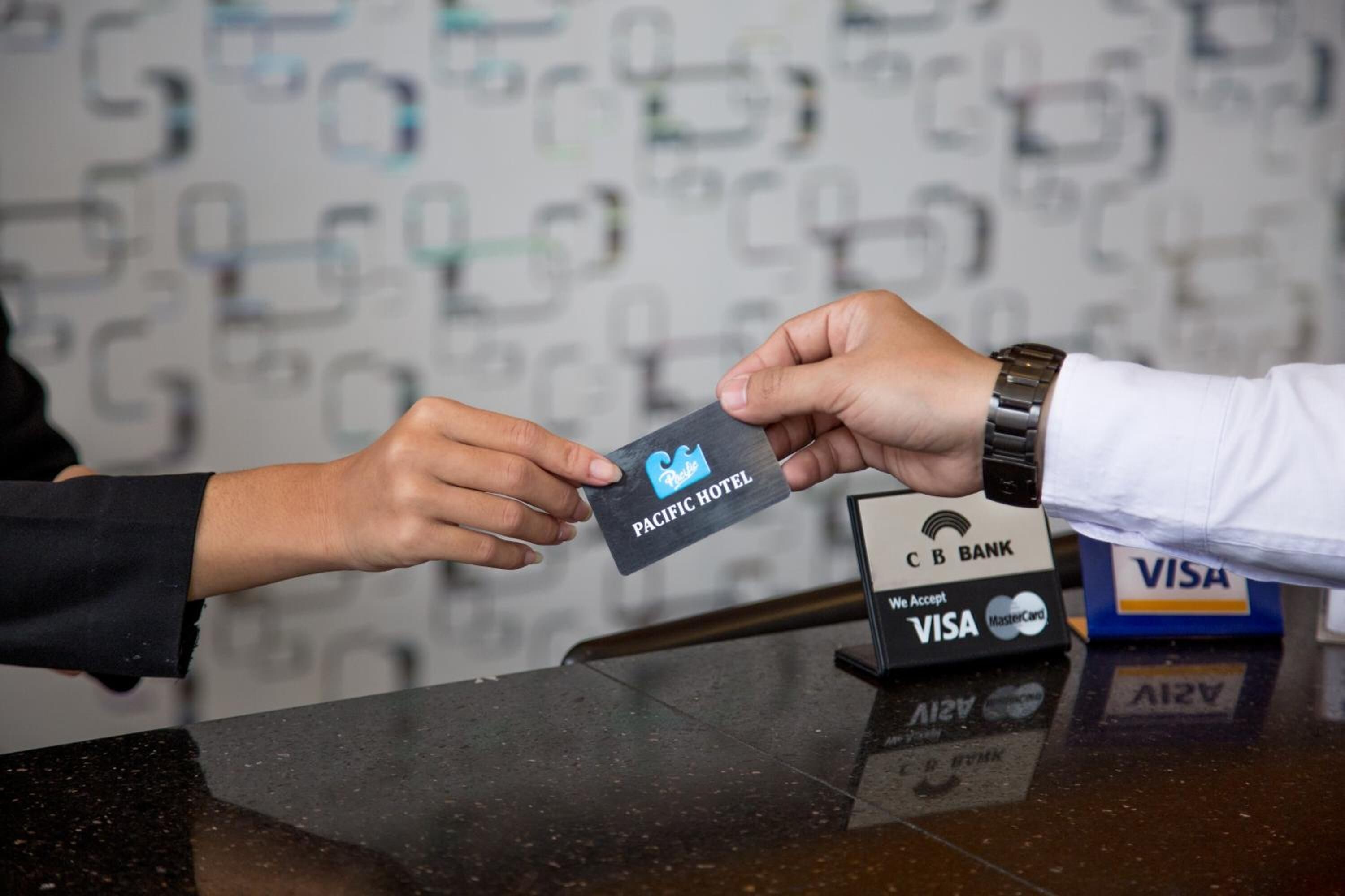 A person handing over a Pacific Hotel card to another person at a reception desk with a C.B. Bank Visa and MasterCard machine.