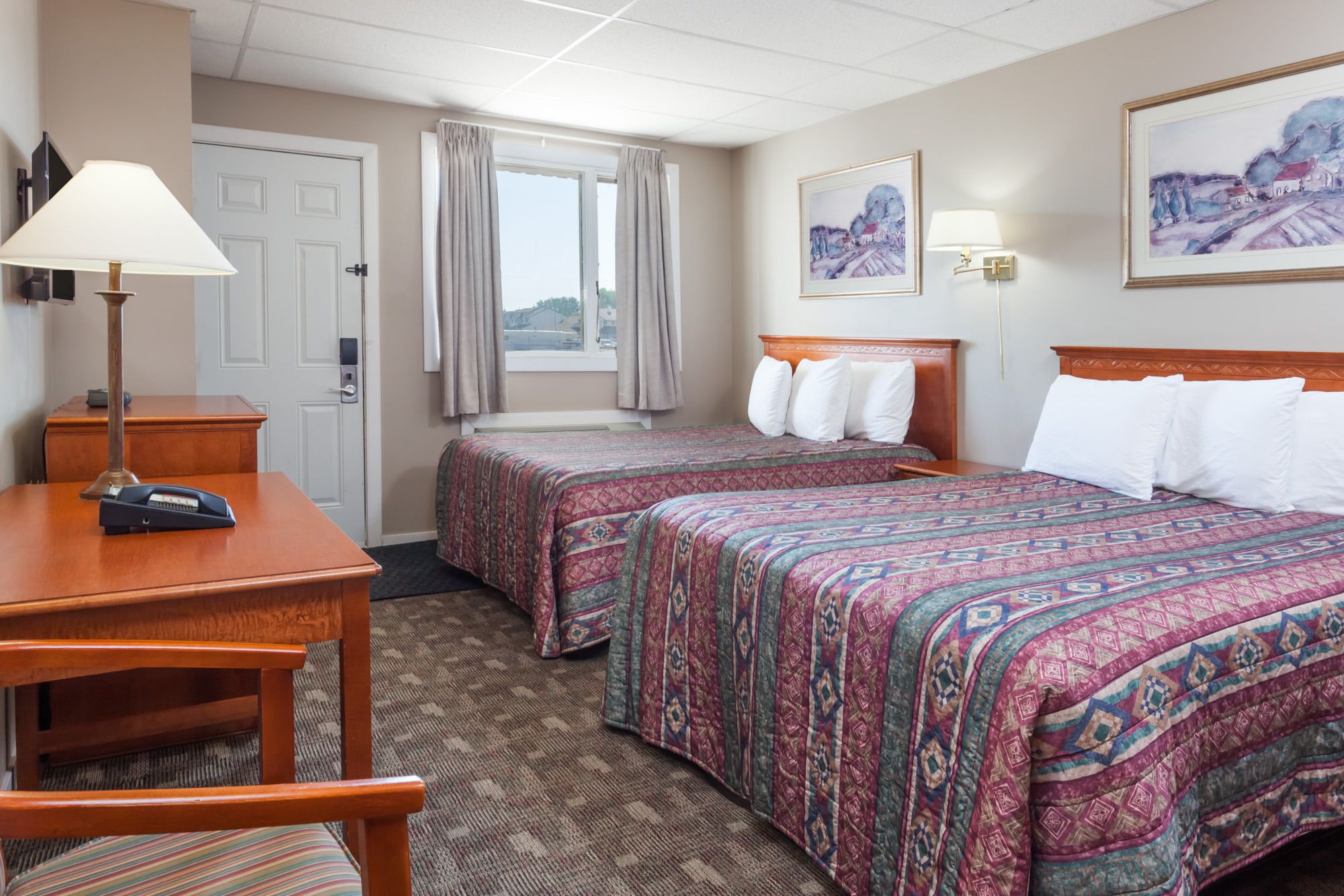 A hotel room with two beds, a desk, a chair, a lamp, and a window with curtains.