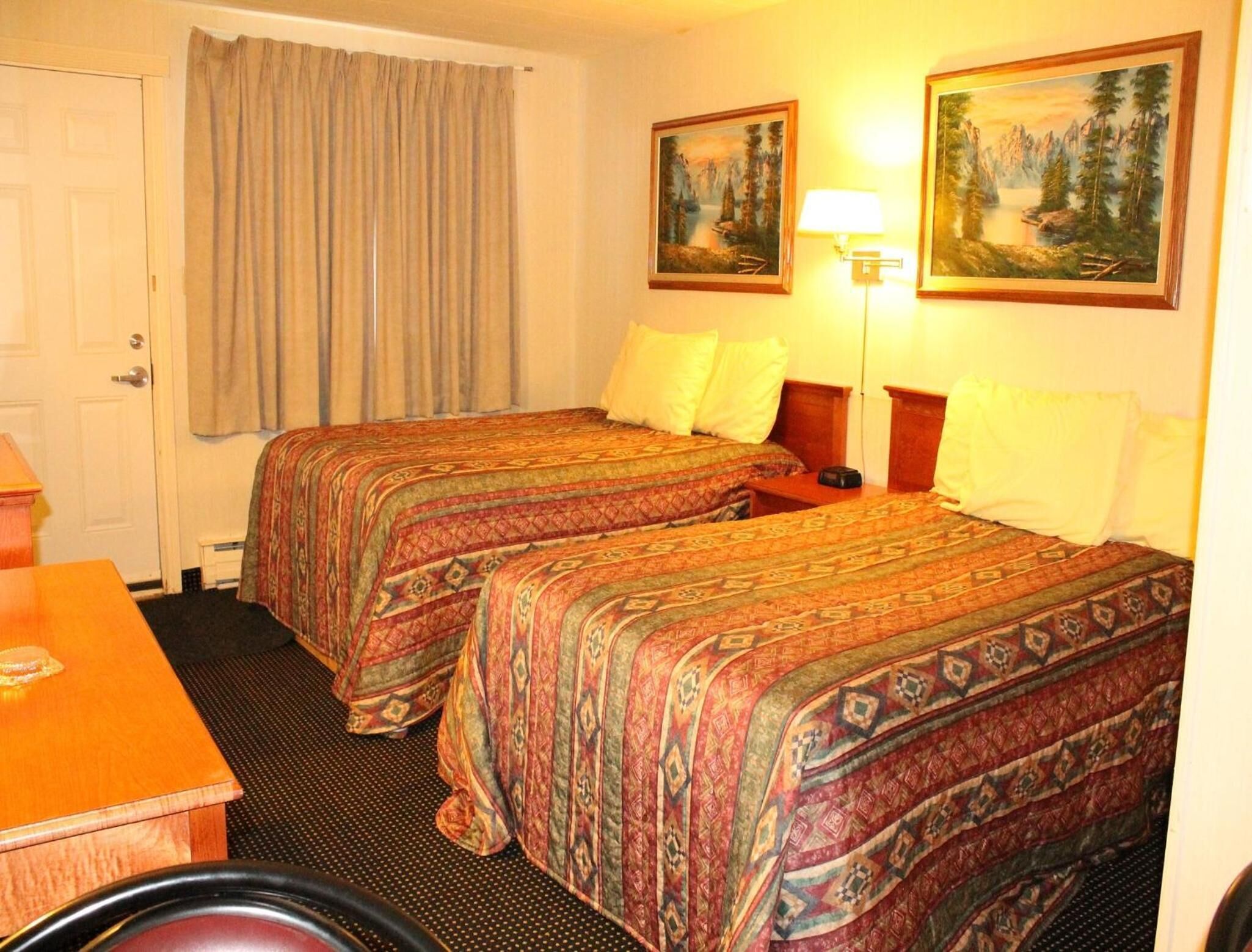 A hotel room with two beds, a desk, a chair, and two paintings on the wall.