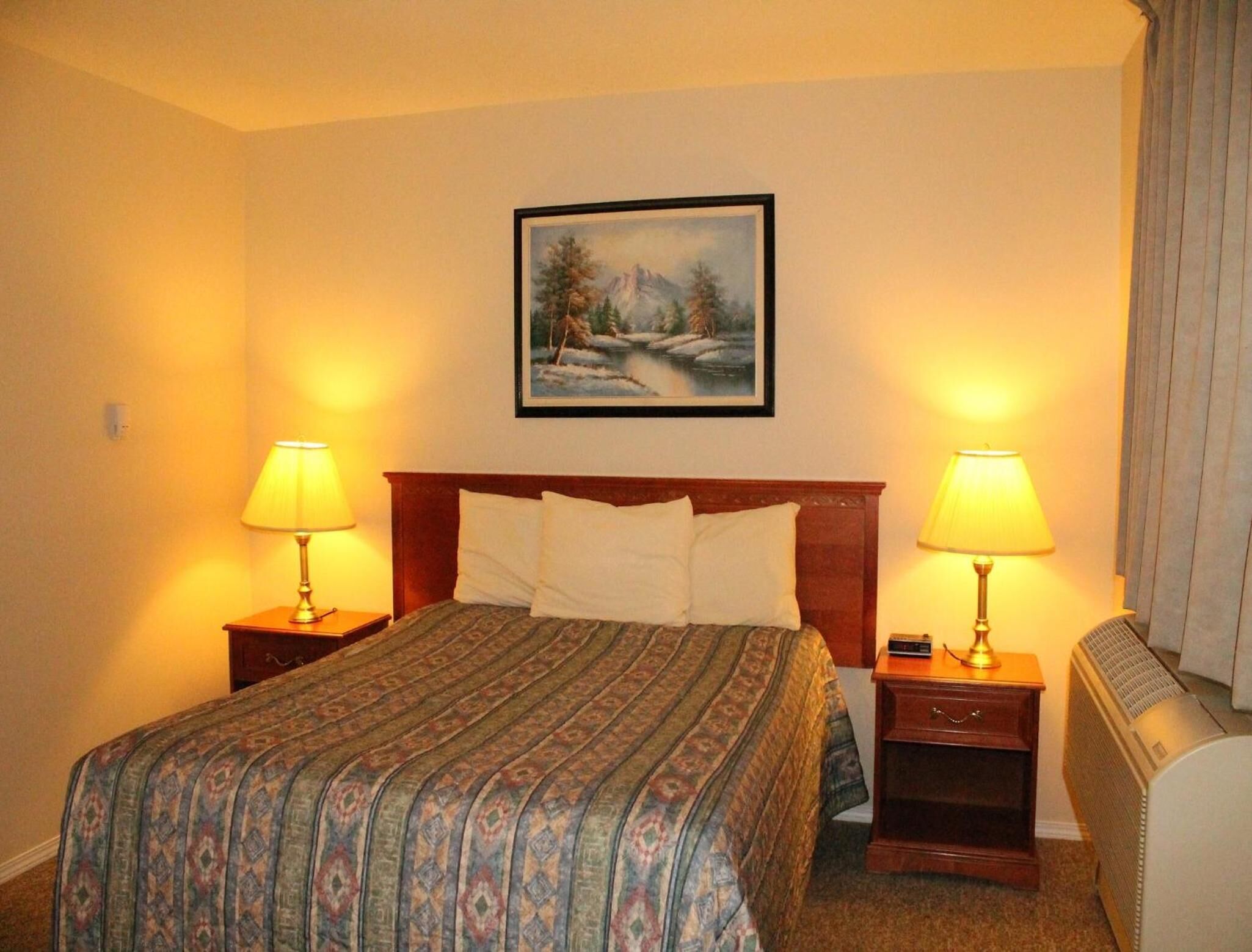 A hotel room with a bed, two bedside lamps, a painting on the wall, and a radiator.