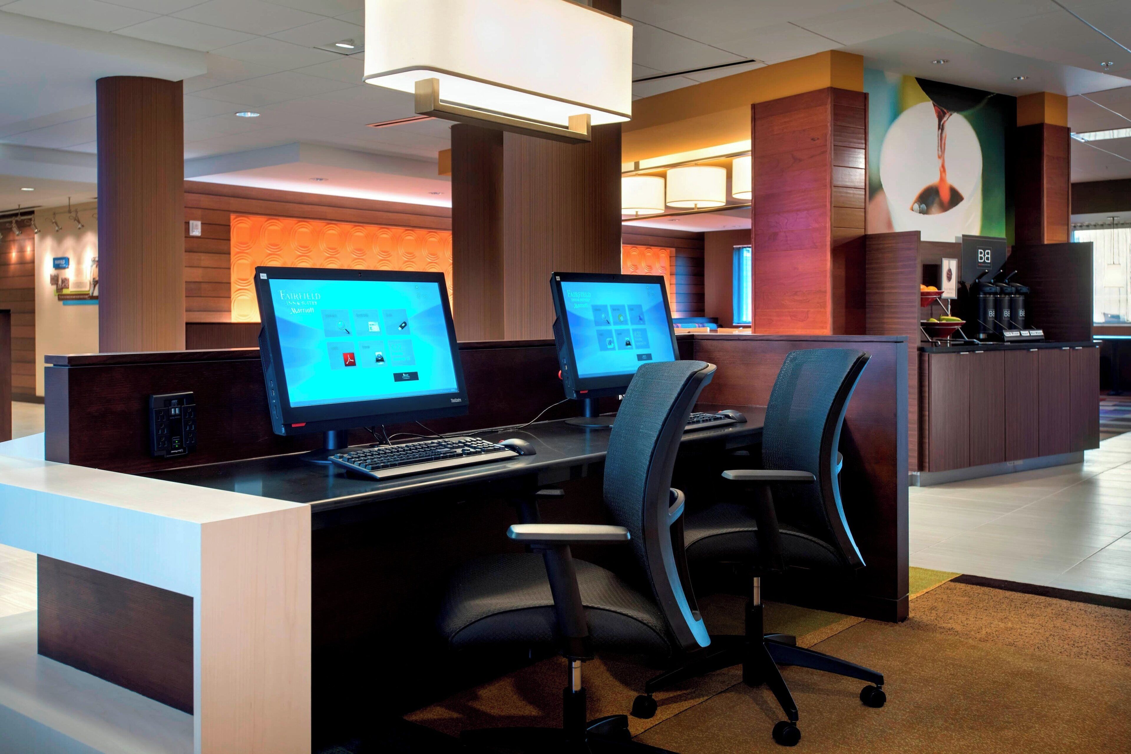A modern reception desk with two computer monitors, a coffee machine, and a seating area with chairs.