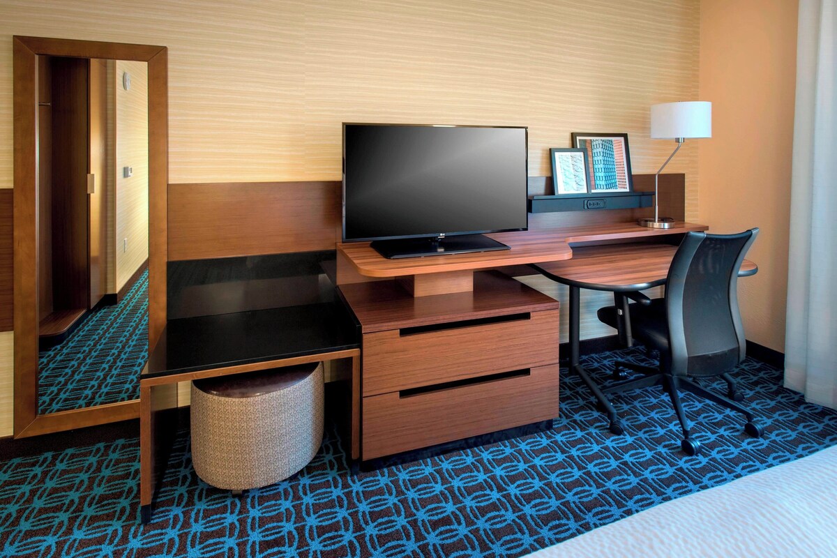 A hotel room with a wooden desk, a flat-screen TV, a chair, a lamp, and a waste basket.