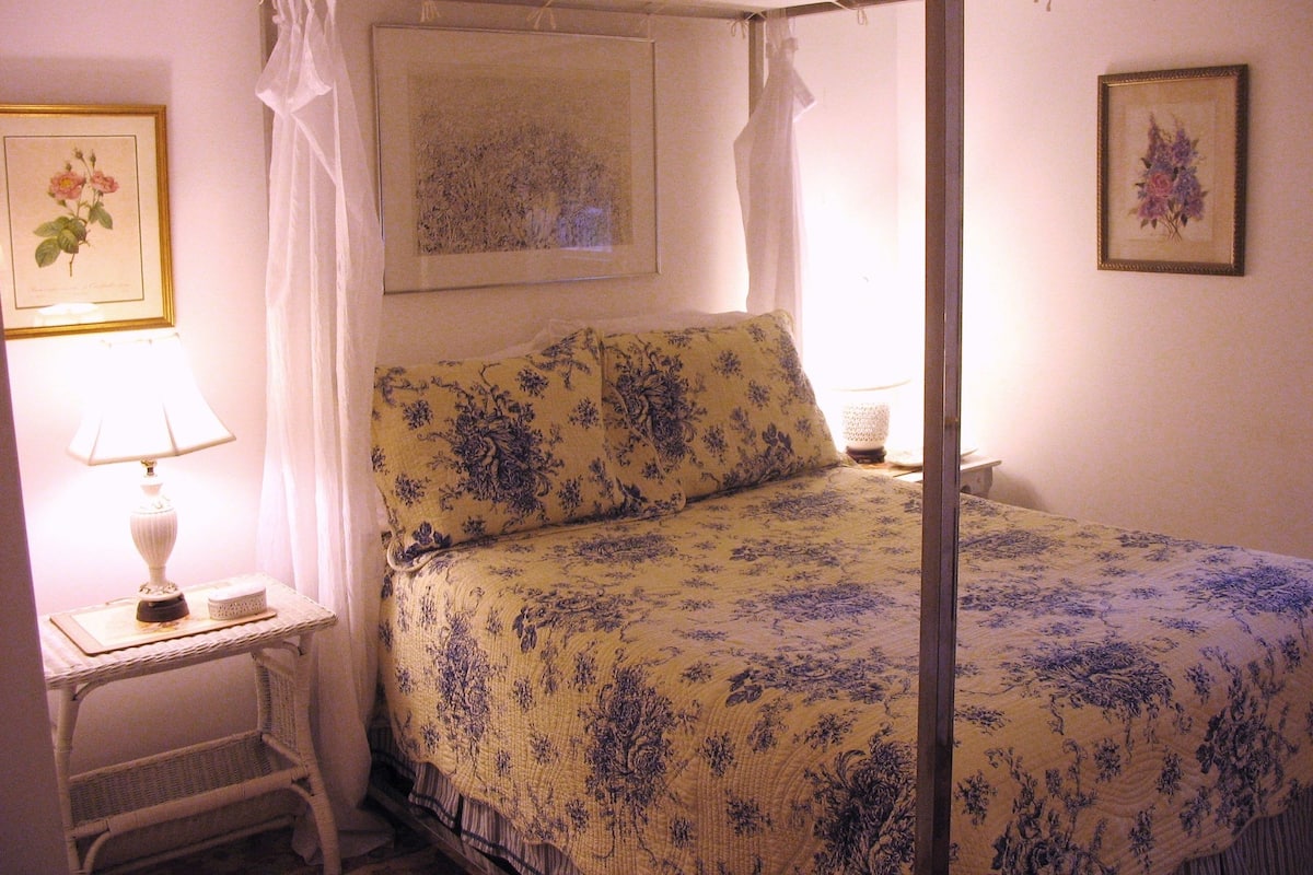 A four-poster canopy bed with floral bedding, a bedside table with a lamp, and framed floral artwork on the walls.