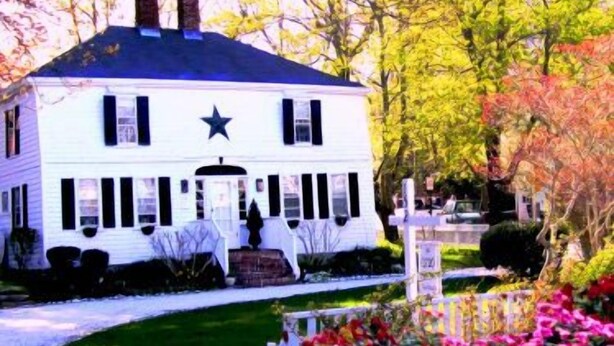 A white house with a star on the front, surrounded by a well-maintained garden with flowers and a picket fence. - The 1720 House (Vineyard Haven)