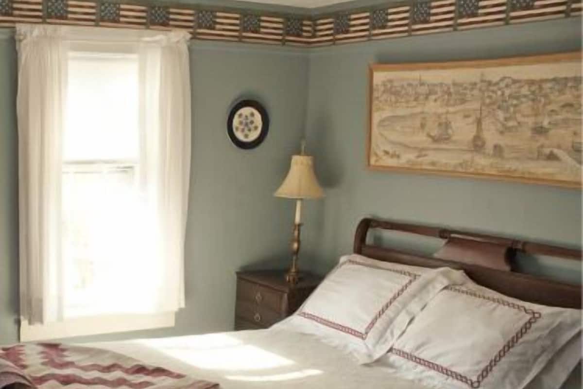 A traditional bedroom with a wooden bed, a nightstand with a lamp, a window with curtains, and a wall hanging.