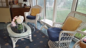 A cozy room with wicker furniture, a glass table with a vase of flowers, and a view through large windows. - The 1720 House (Vineyard Haven)