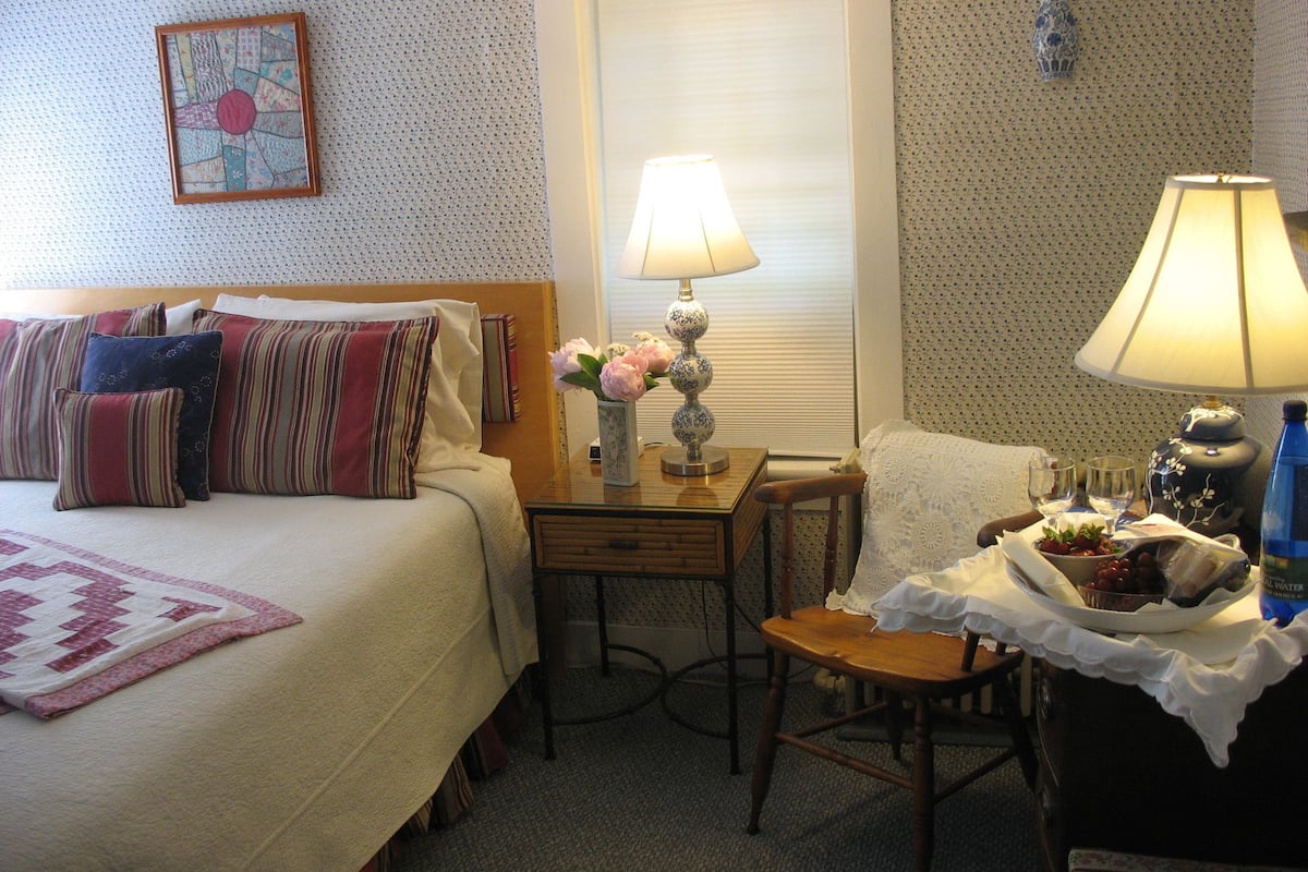 A hotel room with a bed, a chair, a table with a lamp, and a vase with flowers.