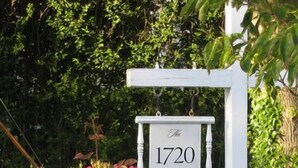A white sign with the text "The 1720 House" hanging above a white fence, surrounded by lush green foliage and pink roses. - The 1720 House (Vineyard Haven)