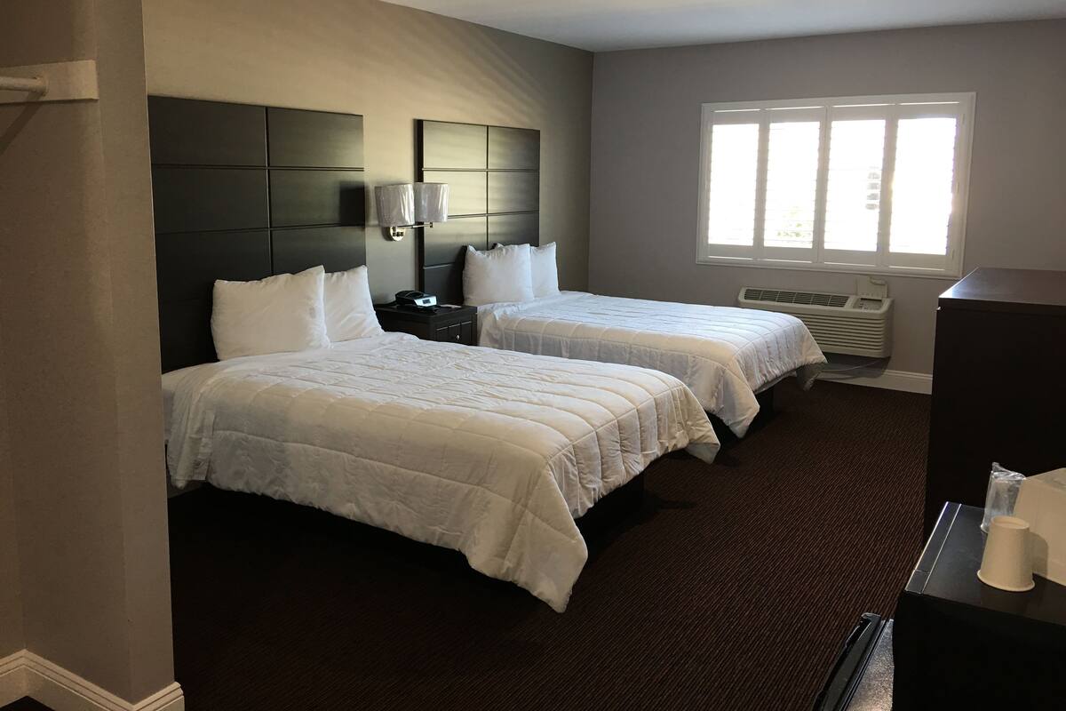 A hotel room with two beds, a dark headboard, a window with blinds, and a desk with a cup.