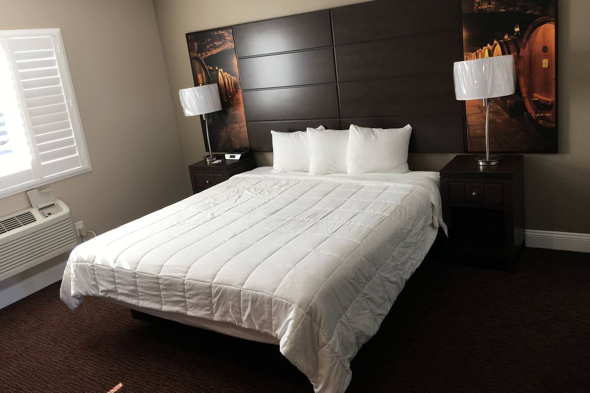 A hotel room with a large bed, two bedside tables with lamps, a wall-mounted artwork, and a window with blinds.