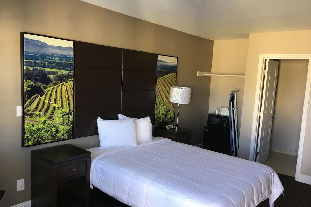 A hotel room with a large bed, a nightstand, a lamp, a microwave, and a door.