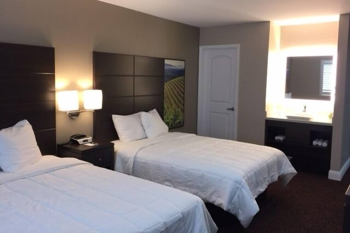 A hotel room with two beds, a nightstand, a lamp, a painting, a door, and a bathroom with a mirror and sink.