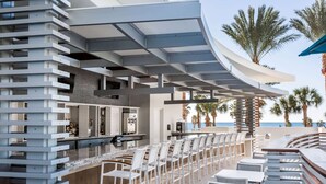 Zona de bar moderna al aire libre con techo curvo, taburetes blancos y vista al océano.