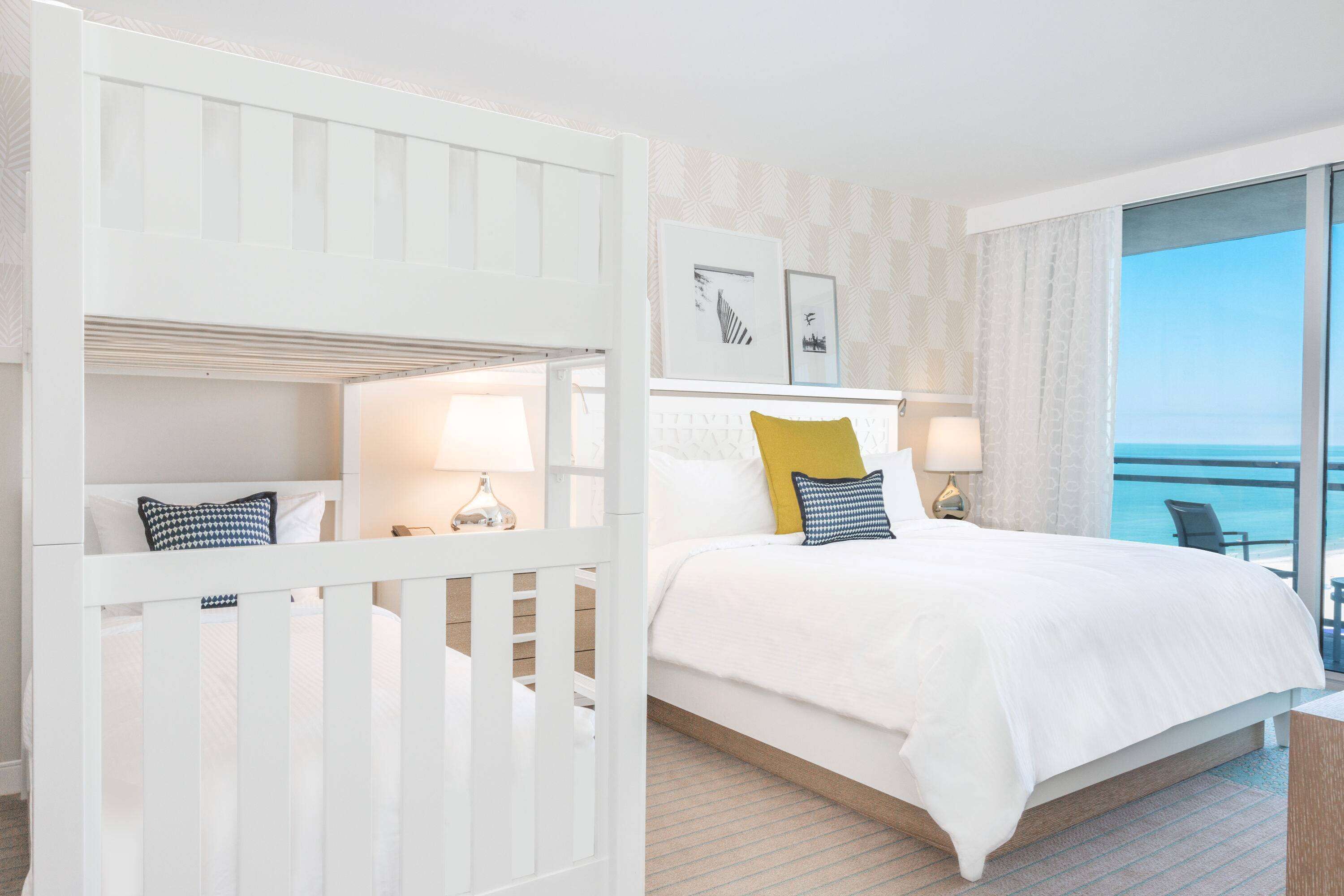A hotel room with a bed, a crib, a balcony with a view of the sea, and a small table with a lamp.