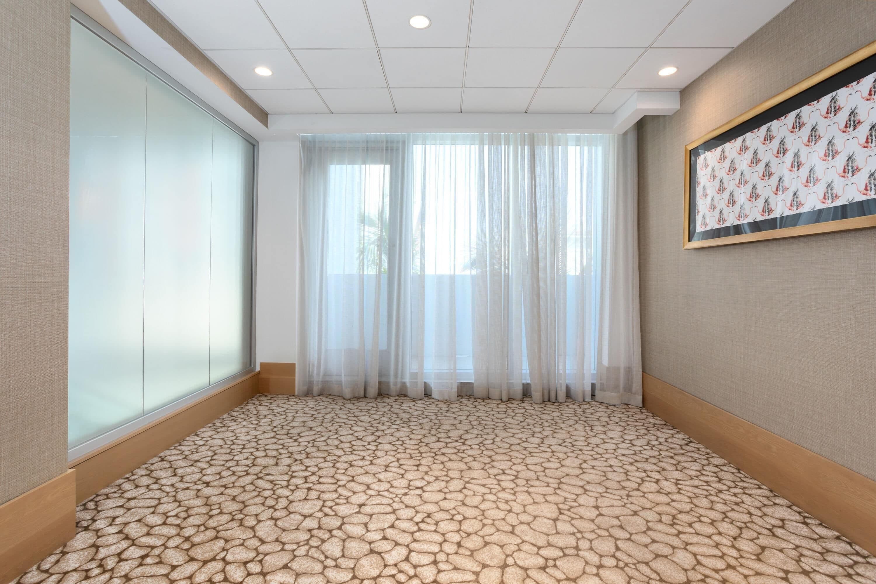 A room with a patterned floor, large windows with sheer curtains, and a framed picture on the wall.