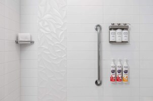Un baño con azulejos blancos, un perchero, una barra de apoyo y tres botellas de champú.