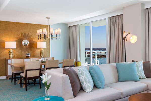 Habitación de hotel con zona de comedor, sofá con cojines estampados, un espejo grande y vistas a la ciudad.