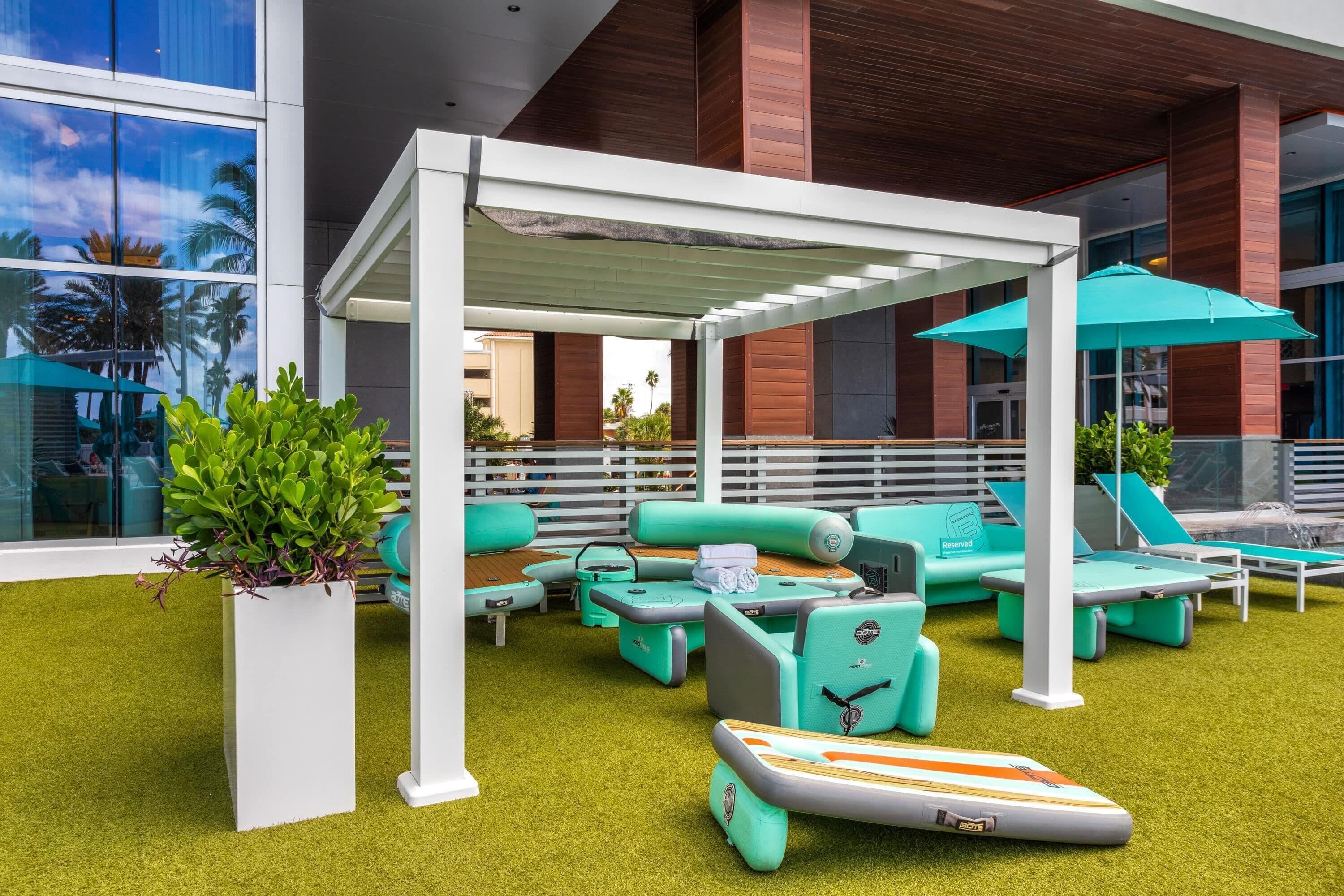 Un espace de détente extérieur moderne, avec une pelouse artificielle verte, des sièges blancs et turquoise, et une zone abritée par des parasols.