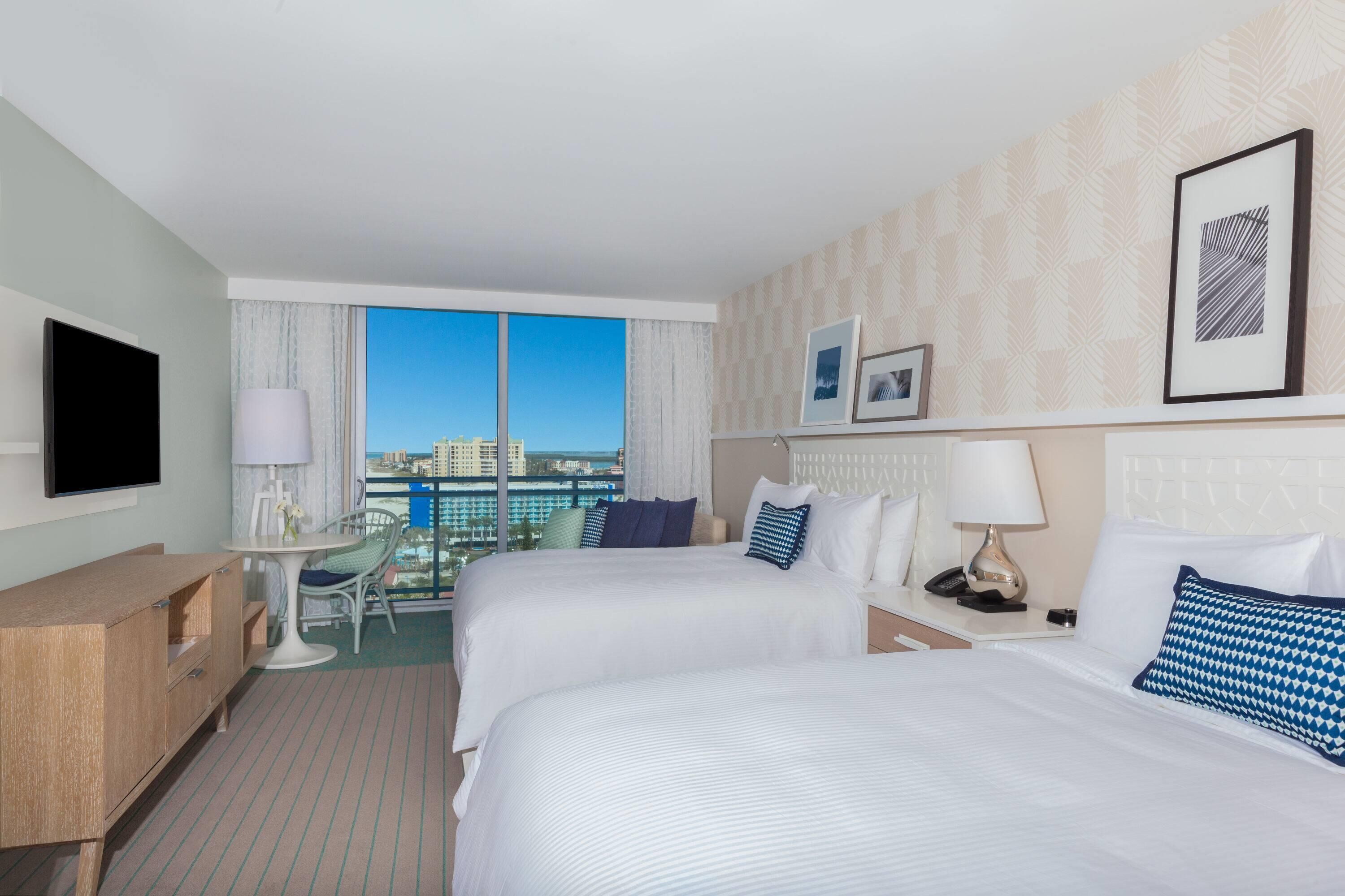 A hotel room with two beds, a desk, a chair, a TV, and a window with a city view.